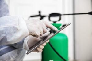 Man in virus protective suit and mask looking and typing on tablet, disinfecting buildings of coronavirus with the sprayer. Infection prevention and control of epidemic. World pandemic.